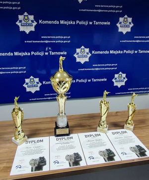 Puchary oraz dyplomy na tle niebieskiej ścianki z logo i napisem Komenda Miejska Policji w Tarnowie
