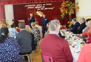spotkanie z seniorami w trakcie którego dwóch policjantów stoi przed zgromadzonymi seniorami a jeden z nich trzyma mikrofon i informuje o zagrożeniach
