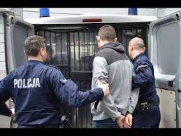 Dwóch Policjantów  prowadzi zatrzymaną osobę, która ma założone kajdanki na ręce trzymane z tyłu do policyjnego radiowozu