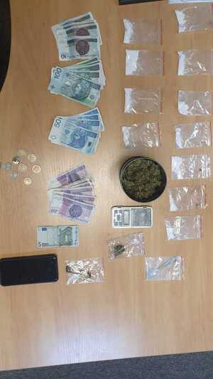 Na biurku rozłożone są waga elektroniczna, banknoty o nominale 10 zł ,20 zł, 50 zł,100 zł, 3 Euro, ciemna puszka z zawartością marihuany, woreczki foliowe z poporcjowaną  zawartością marihuany i amfetaminy.