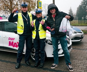 policjant, policjantka i dziennikarz radiowy prezentują odblaski rozdawane przechodniom w tle samochód z logo stacji radiowej