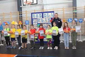 grupa dzieci przedstawicieli poszczególnych drużyn biorących udział w turnieju bezpieczeństwa o ruchu drogowym wraz z policjantem, w tle znaki drogowe i tablica z pracami plastycznymi