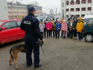 przewodnik psa stoi frontem do dzieci, pies na smyczy. Policjant w ręku trzyma kaganiec. Dzięki ustawione w rzędach na parkingu jednostki
