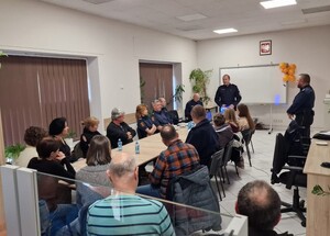 Grupa osób siedząca wokół stołu w tym umundurowani strażnicy miejscy oraz policjant. Ponadto dwaj umundurowani  policjanci stojący obok