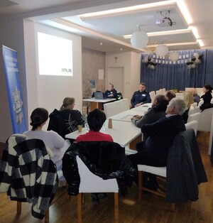 Grupa osób siedzących tyłem wokół stołów, a także dwaj umundurowai policjnaci. Po lewej stronie policyjny baner, a na ścianie wyświetlana prezentacja.