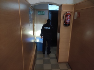 Policjant z latarką sprawdza korytarz z w poszukiwaniu niebezpiecznego urządzenia. Policjant stoi plecami do zdjęcia.