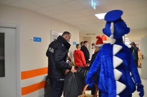 policyjni pomocnicy Świętego Mikołaja z Inspektorem Wawelkiem na idąc szpitalnym korytarzem do dzieci, niosą worki z prezentami.