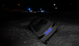 Samochód koloru białego znajdujący się w przydrożnym rowie. W tle dwa policyjne radiowozy z włączonymi sygnałami świetlnymi