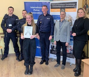 Na zdjęciu widać stojących przodem, trzech umundurowanych policjantów. Obok mężczyznę i kobietę w strojach cywilnych. Przed nimi dziewczyna trzymająca w ręku dyplom