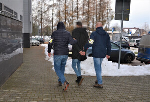 dwóch nieumundurowanych policjantów prowadzi zatrzymanego mężczyznę do radiowozu