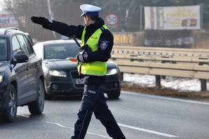 Policjant ruchu drogowego podczas zatrzymywania pojazdu do kontroli, stoi na jezdni, ręką wskazuje miejsce do zatrzymania pojazdu