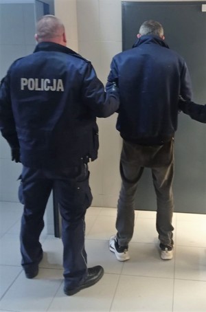 Umundurowany policjant trzymający za ramię zatrzymanego mężczynę, Osoby stoją tyłem na tle drzwi.