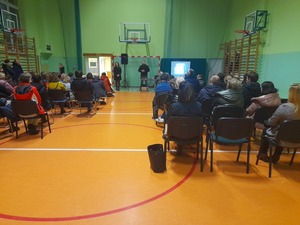 prelekcje na sali gimnastycznej w szkole, widoczna siedząca publiczność rodziców, przodem do niech stoi przed nimi pani z dyrekcji szkoły obok policyjny psycholog po cywilnemu