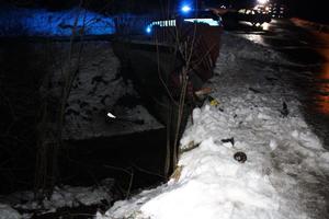 Zaśnieżone pobocze drogi z połamaną barierką mostu nad potkiem. w tle radiowów i samochód biorący udzial w wypadku.