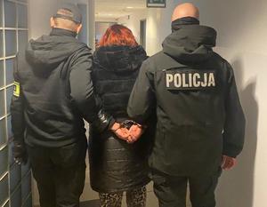 dwóch policjantów prowadzi korytarzem zatrzymaną kobietę z założonymi kajdankami na ręce trzymane z tyłu.