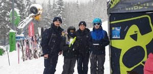 Na stoku narciarkim  od lewej umundurowany policjant, chłopak w stroju narciarkim, umundurowana policjantka i obok niej chłopak w stroju narciarkim. W tle poukładane stojąco narty i ośnieżone drzewa.