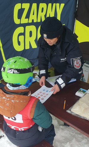 Umundurowana policjantka pokazująca na kartce znaki drogowe dziecku ubranemu w strój narciarki i kask. Dziecko siedzi tyłem. W tle napis Czarny Groń.