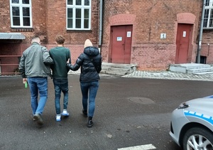 dwoje polcijantów kryminalnych prowadzi osobę zatrzymaną przez dziedziniec komendy