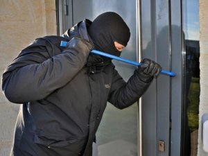 Zamaskowany mężczyzna w czarnej kurtce i kominiarce łomem podważa drzwi
