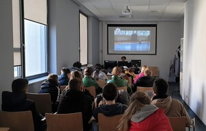 Uczniowie siedzący tyłem na krzesłach w pomieszczeniu komendy. Przed nimi za biurkiem siedzi umundurowana policjantka. Na ścianie projektor, na którym wyświetlana jest prezentacja