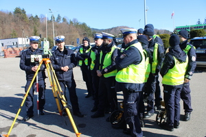 Grupa policjantów ruchu drogowego omawają zasadę pracy urządzenia do laserowego pomiaru punktów przy sporządzaniu planu sytuacyjnego podczas zdarzenia drogowego