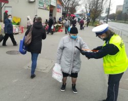policjantka ruchu drogowego wręcza starszej kobiecie opaskę odblaskową, w tle budynki placu targowego oraz ludzie.jpg