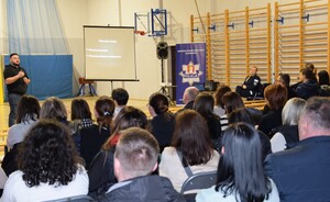 część rodziców siedzących tyłem na sali gimnastycznej przed nimi psycholog policyjny prowadzący prelekcje z wyświetloną prezentacją, obok siedząca policjantka