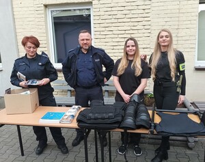 Cztery osoby stojące przodem, od lewej umundurowana policjnatka i policjnt orz dwie dziewczyny ubrane na czarno. Ostatnia na przedramieniu ma założoną opaskę z napoisem Policja. Na stoliku sprzęt policyjny.