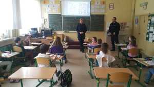 Policjantka i policjant w klasie prowadza zajęci z dziećmi. Dzieci siedzą w szkolnych ławkach na wprost policjantów.