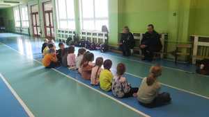 Policjantka i policjant siedzą na ławeczce przodem do dzieci i rozmawiają z nimi o bezpieczeństwie. Dzieci siedzą na podłodze, przodem do policjantów  w sali gimnastycznej.