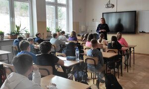 Policjantka na spotkaniu z uczniami. Dzieci siedzą w szkolnych ławkach twarzą do policjantki. Zdjęcie w sali szkolnej.