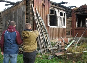 Na zdjęciu widać dwie osoby po lewej stronie policjant w ubiorze cywilnym ( granatowo-bordowa kurtka) po prawej stronie podejrzany w kurtce w kolorze khaki. Mężczyźni stoją przed spalonym pomieszczeniem gospodarczym.