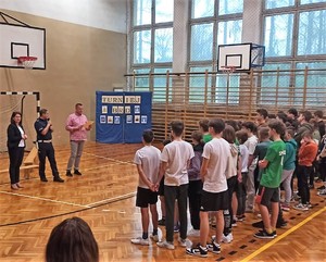 Grupa dzieci stojących tyłem. Przed nimi umundurowany policjant oraz kobieta i mężczyzn w stroju cywilnym. Stoją na sali gimnastycznej.