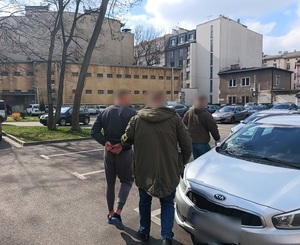 Zatrzymany mężczyzna prowadzony przez funkcjonariuszy przez policyjny parking do radiowozu