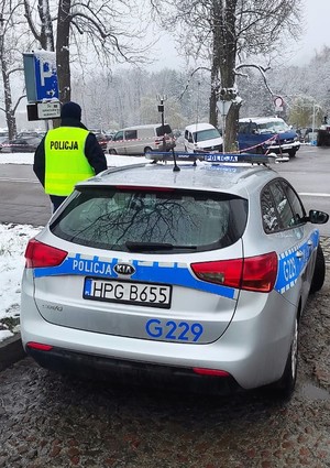Na zdjęciu widać oznakowany radiowóz i stojącego obok niego umundurowanego policjanta w kamizelce odblaskowej z napisem Policja. W tle zaparkowane samochody.