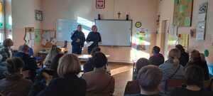 Dzielnicowy i policjantka na spotkaniu z rdzicami. Zajęcia odbywają sie w klasie. Rodzice siedzą w szkolnych ławkach,  przodem do policjantów