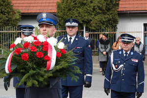 Generał z zastępcami podchodzi do krzyża przed nim maszeruje policjant z wiązanką kwiatów