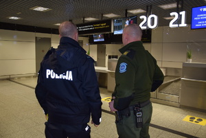 policjant i pracownik Służby Ochrony Lotniska stojący tylem do zdjecia przed nimi strefa bagażowa i odpraw