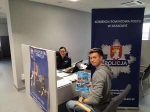policjantka siedząca przy biurku w urzędzie pracy, przed nią mężczyzna trzymajacy w ręku ulotkę, obok wywieszony plakat wstąp do Policji, z drugiej strony baner KPP w Krakowie