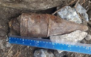 Zbliżenie na głowicę przeciwpacenrną leżąca na ziemi, obok przymiar liniowy wskazujący na wługość pocisku około 25 cm