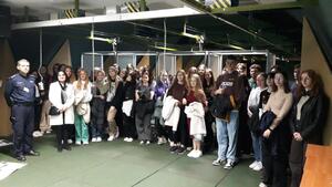 Młodzież siedząca na sali słucha informacji pracownika Policji na temat warunków przyjęcia do służby w Polcji, obok stoi policjnat, na stole leży otwarta walizka technika kryminalistyki i kombinezon