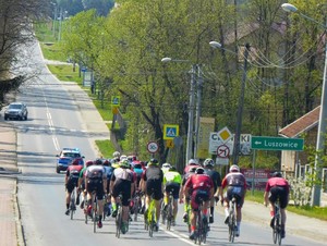 peleton rowerzystów poruszający się jezdnią, przed nimi radiowóz oznakowany. Obok jezdni znak kierujący do miejscowości Luszowice