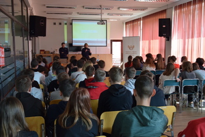 Policjnatka i policjant w sali olkuskiej biblioteki prowadzą szkolenie dla uczniów klas 8. Młodziez siedzi w rzędach. W tle wyswietlony slajd prezentacji.