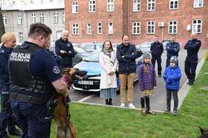 przewodnik psa opowiada o swojej pracy dzieciom i ch rodzicom. obok policjanta jest pies, a w tle policjanci