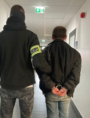 Po lewej stronie policjant w stroju cywilnym, na ręce ma założoną opaskę odblaskową z napism Policja. Trzyma za rękę zatrzymanego mężcyznę, który ma założone kajdanki na rękach z tyłu.