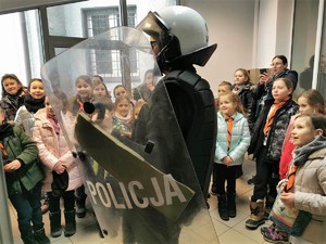 Na zdjęciu widać osobę ubraną w kask policyjny, kamizelkę kuloodporną, w ręku trzyma tarczę ochronną z napisem Policja. Wokoło grupa dzieci