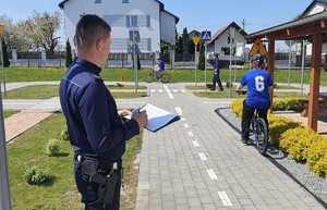 Na zdjęciu widać dwóch umundurowanych policjantów którzy oceniają jazdę dwóch osób na rowerze  po specjalnie przygotowanym torze ze znakami drogowymi. W tle domy i smochód.