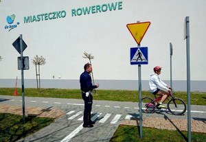 Umundurowany policjant ocenia chłopca jadącego na rowerze po specjalnie przygotowanym torze ze znakami drogowymi. Na ścianie budynku napis Miasteczko Rowerowe