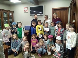 Grupa dzieci stojących przodem. Za nimi dwie kobiety w stroju cywilnym i umundurowana policjantka. Na ścianie Godło Polski , telewizor.