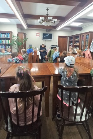 W pomieszczeniu biblioteki grupa dzieci siedzących na krzesłach przy stole. Pośrodku umundurowana policjantka siedząca na krześle, która czyta ksiązkę. W tle regały z książkami, telewizor, drzwi.
